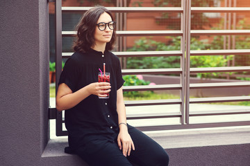 Young stylish woman in glasses with lemonade, outdoors. Girl in casual clothes, holding summer cocktail at cafe terrace, summertime. Woman with glass of refreshment drink in sunny day.