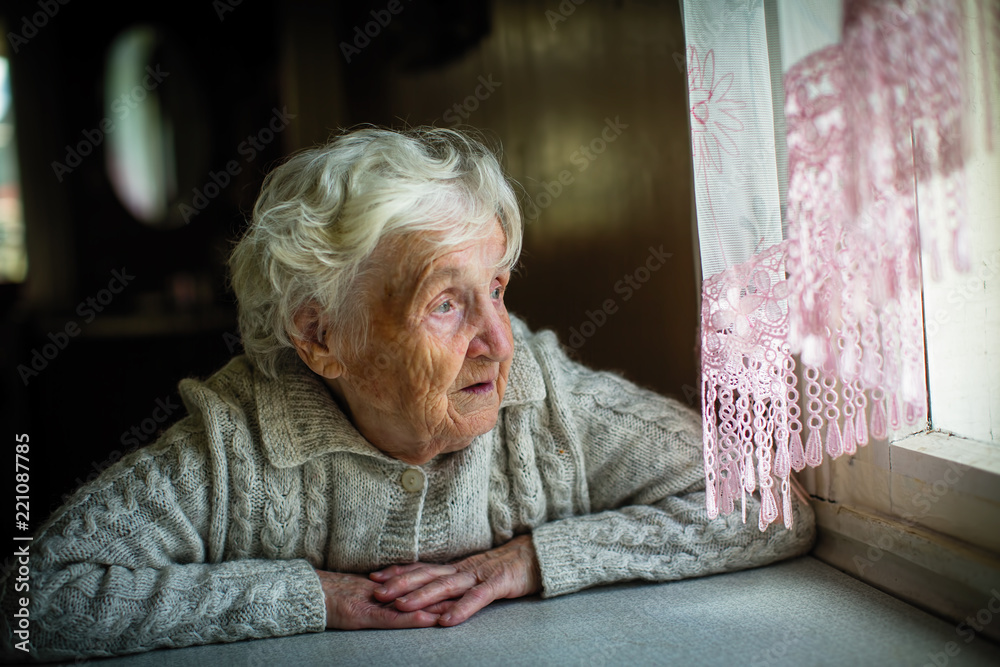 Wall mural Gray-haired elderly woman sits and looks out the window.