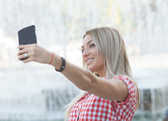 Carefree and happy, sunny summer mood. Charming young lady is making selfie on a camera. She is wearing formal wear, smiling, while on a walk in town outdoors