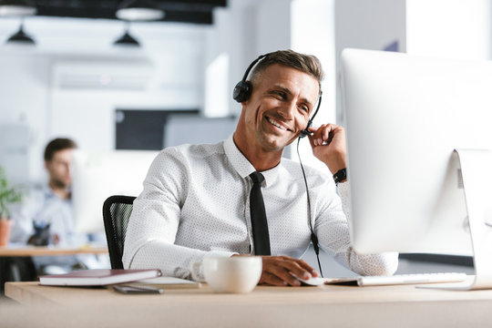 Photo of smiling operator man 30s wearing office clothes and headset, sitting by computer in call center
