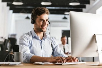 Photo of businesslike man 20s wearing office clothes and headset, working on computer in call center - obrazy, fototapety, plakaty