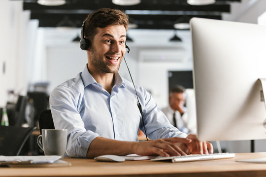 Photo of happy operator man 20s wearing office clothes and headset, sitting by computer in call center