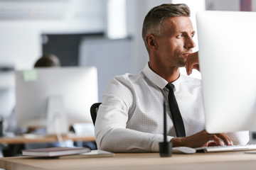 Naklejka premium Image of adult businessman 30s wearing white shirt and tie, sitting by computer in office