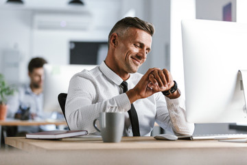 Image of adult businessman 30s wearing white shirt and tie sitting at desk in office by computer,...