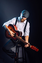 A bearded man in suit and hat sitting and playing on the acoustic guitar. Studio portrait