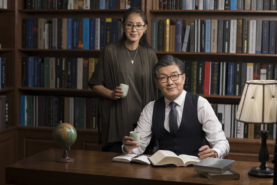 Cheerful mature couple reading book and drinking tea in study