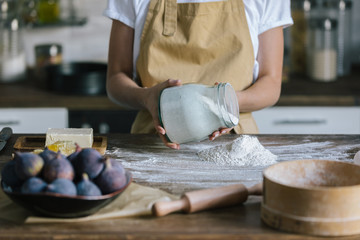 cropped shot of woman pouring flour on rustic wooden table with fig pie ingredients