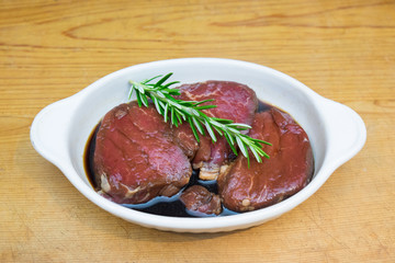 Meat marinating: Beef steak eye fillet in white dish of soy sauce marinade and rosemary herb sprig on wooden background with copy space