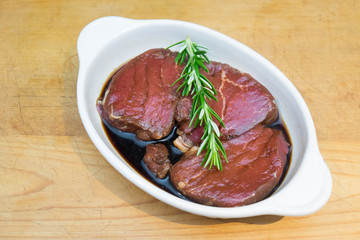 Meat marinating: Beef steak eye fillet in white dish of soy sauce marinade and rosemary herb sprig on wooden background - diagonal composition