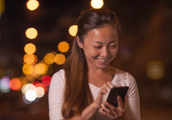 Portrait of beautiful young asian girl texting on cell phone outdoor over blurred night street background, selective focus. iGen mobile style of life.