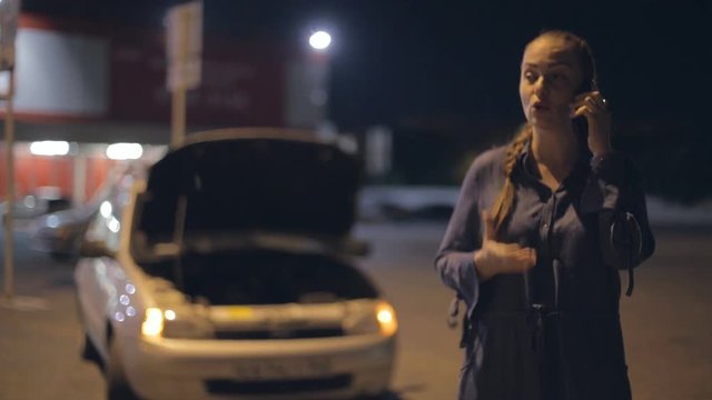 a young woman standing near a broken car at night on the road in the rain, calling on the phone, crying and swearing, asking for help.