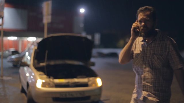 a man stands in the rain at night near a broken car, calls on the phone, swears and asks for help.