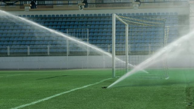 Water jets sprinkling on green grass of soccer stadium. Sports field watering irrigation at daylight. Empty sports stadium with tribune and soccer goal on field. Water sprinkler at football field