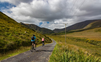 Fototapeta na wymiar Active couple biking on a rural country road in the scenic mountain valley landscape of the Dingle peninsula, County Kerry in the Republic of Ireland