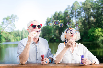 Grandfather and grandmother. Grandfather and grandmother feeling entertained while using soap bubbles of their grandchildren - obrazy, fototapety, plakaty