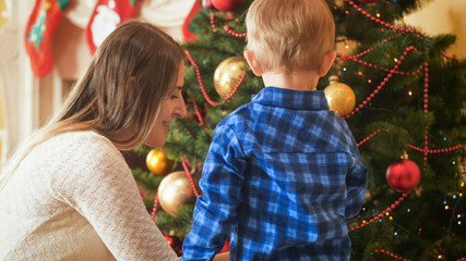 Fototapeta na wymiar Portrait of young mother helping her little son decorating Christmas tree in living room