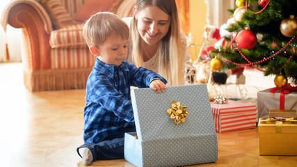 Cheerful little boy opening Christmas gift at morning under Christmas tree