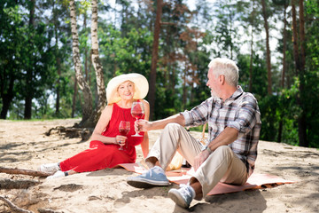 Lady in hat. Beaming elderly lady wearing summer hat and long red dress holding glass of red wine sitting near husband