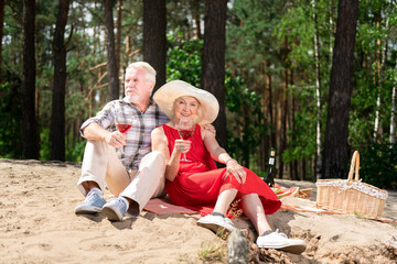 Fototapeta premium Nice picnic. Cute elderly wife and husband wearing sneakers having picnic drinking glasses of red wine
