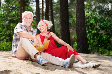 Lady with apple. Smiling elderly lady eating little red apple while sitting on sand near her bearded husband