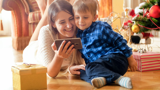 Portrait of cheerful smiling mother with baby lying on floor at living room and watching video on smart phone