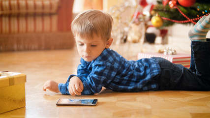 Cute 3 years old toddler boy lying under Christmas tree and watching cartoons on mobile phone