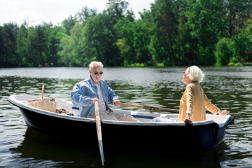 Fototapeta na wymiar Paddles and boat. Happy elderly man holding paddles while sitting in boat with his appealing stylish wife
