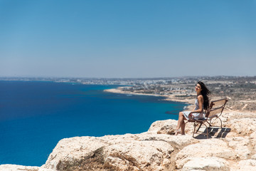Brunette in a dress sits on a bench on the edge of a cliff overlooking the sea