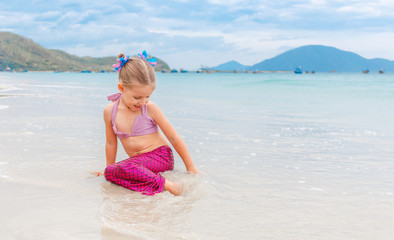 Little beautiful girl dressed in swimsuit as a mermaid sits on the seashore