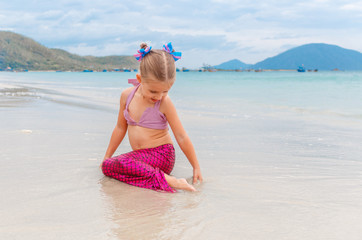 Little beautiful girl dressed in swimsuit as a mermaid sits on the seashore