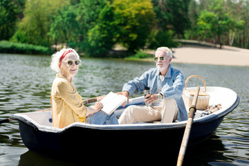 Fototapeta na wymiar Reading time. Happy elderly woman wearing sunglasses reading book during amazing boat ride with her man