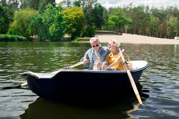 Fototapeta na wymiar Joyful communication. Couple of beaming pensioners wearing sunglasses enjoying their communication sitting in boat