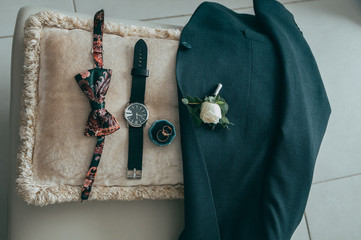 Groom's accessories: a watch, a green jacket, a buttonhole, a butterfly and rings on a tinted beige cushion.