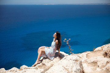 Brunette in a dress sits on the edge of the cliff overlooking the sea