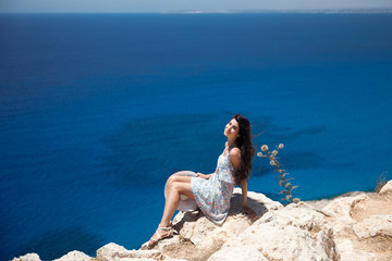 Brunette in a dress sits on the edge of the cliff overlooking the sea