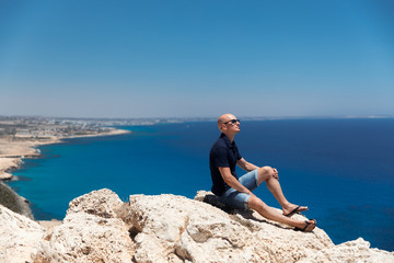 Fototapeta na wymiar Bald man in a T-shirt and shorts sitting alone on top of a mountain overlooking the sea