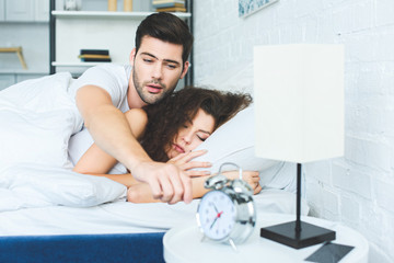 sleepy young man reaching for alarm clock while sleeping with girlfriend in bed