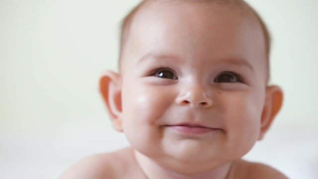 A cute little baby is looking into the camera and is happy on a white bed sheet smiles close up portrait