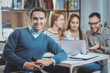 Team cooperation. Cheerful male colleague holding phone and looking at camera