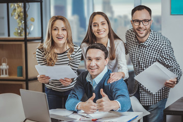 Working atmosphere. Optimistic four colleagues staring at camera and man demonstrating thumb