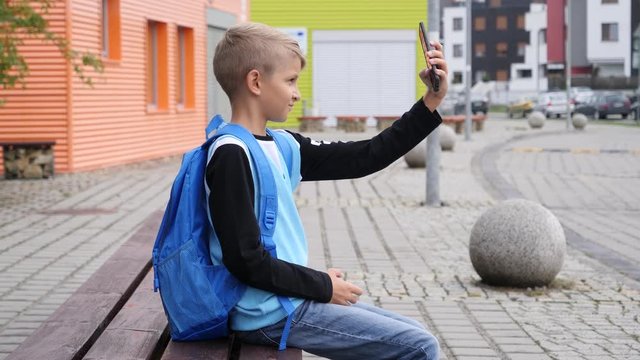 Alone kid boy sit on a bench by a school building taking selfie pictures via smart phone mobile