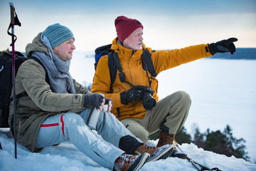 Fototapeta na wymiar Two mature men exploring Finland in winter. Hikers sit on top of rock, take pictures with camera, drink hot coffee from thermos flask. Northern landscape with frozen Baltic Sea and snowy islands.