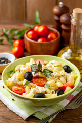 Pasta salad with cherry tomatoes, black olives, feta cheese and basil on wooden background