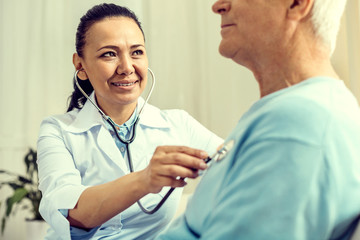 Everything is in norm. Selective focus on a friendly looking nurse smiling while looking at a retired man and checking his heart beating with a stethoscope.
