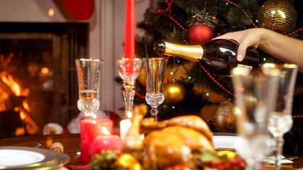 Fototapeta na wymiar Closeup image of filling glasses with champagne on family dinner next to Christmas tree and burning fireplace