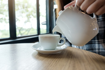 A hand holding teapot and pouring tea into a white cup on wooden table