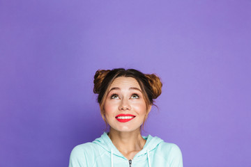 Portrait of cute cheerful girl with two buns looking upward on copyspace and twisting mouth, isolated over violet background in studio