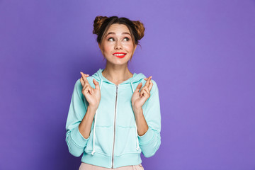 Portrait of funny young girl with two buns keeping fingers crossed and dreaming for good luck, isolated over violet background in studio