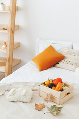 Home decor. Cozy fall bedroom interior: white wall, wooden rack, bed with white linen, light beige plaid, orange pillows,  wooden box with pumpkins, dry autumn leaves, sweater. Autumn decoration.