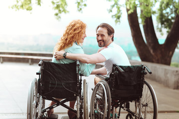 Love her so much. Radiant guy grinning broadly into the camera and hugging his girlfriend while both sitting in their wheelchairs outdoors.